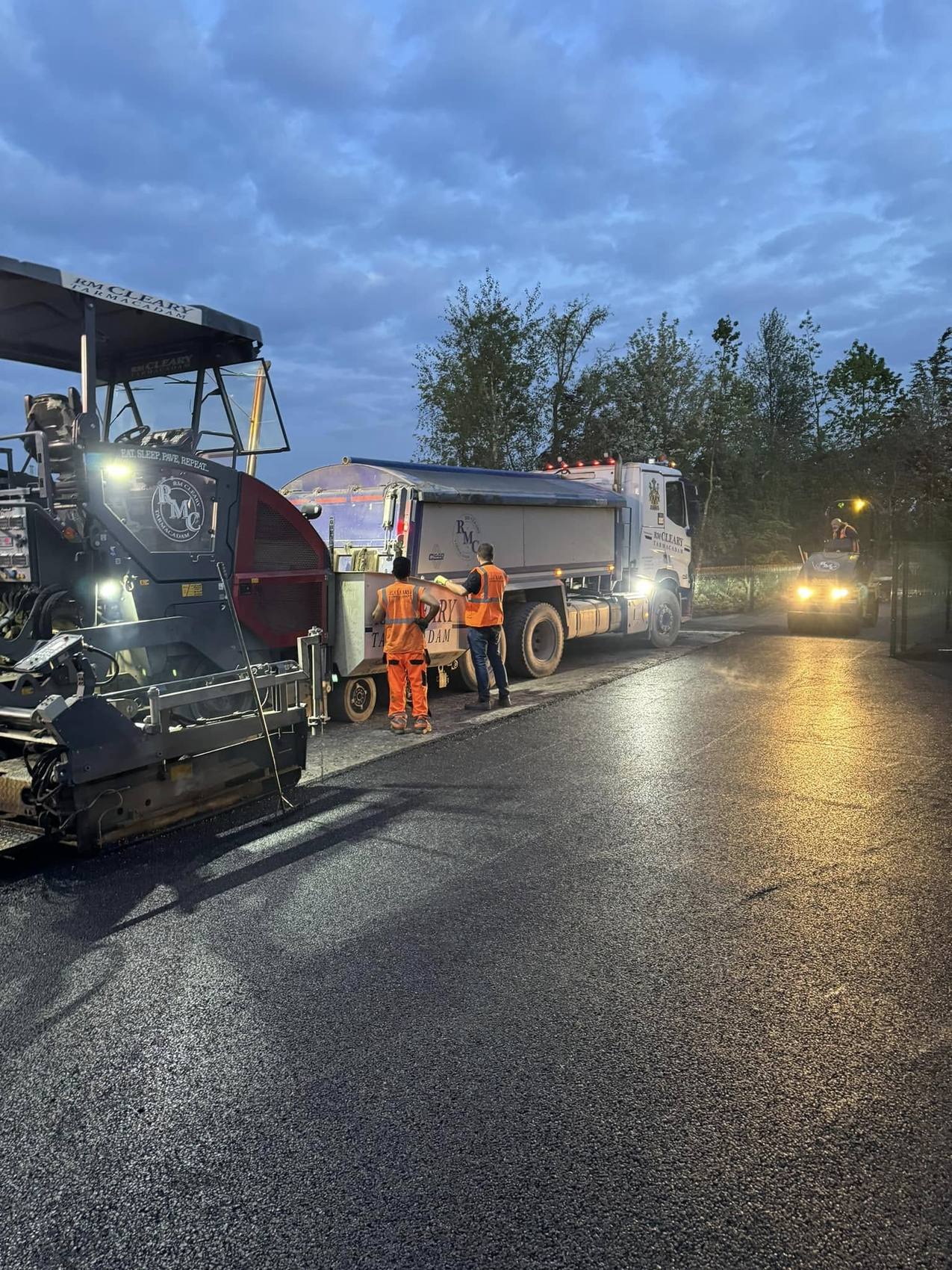 An RM Cleary paver and tanker working at dusk under lights on a resurfacing job.