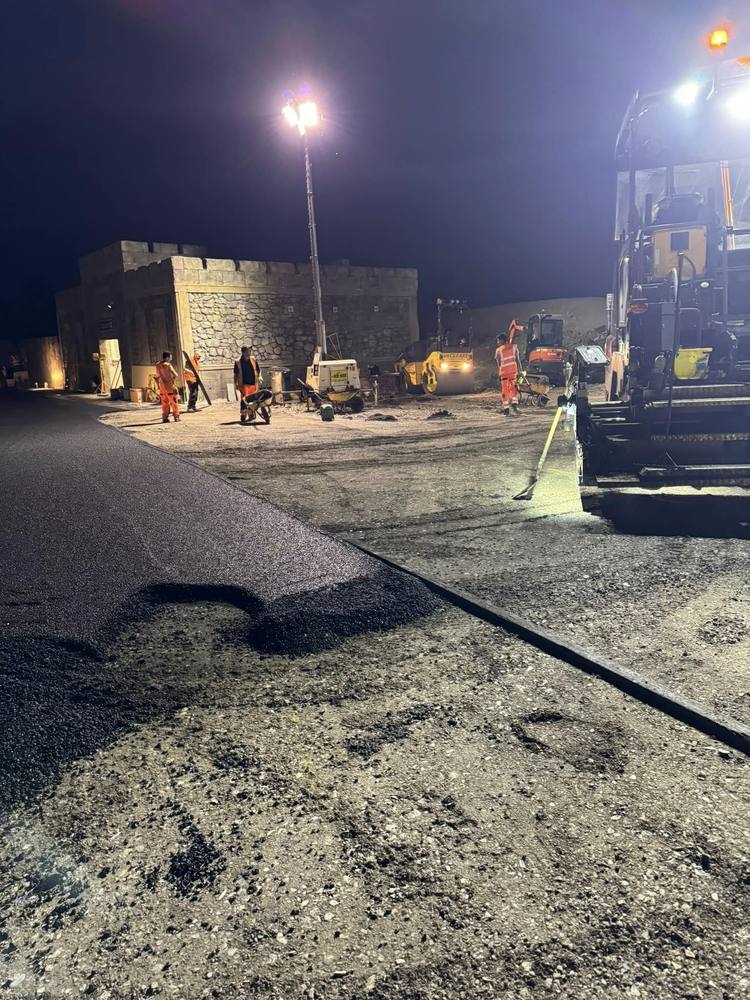 Night works under a floodlight, with the paver laying and a roller following behind.