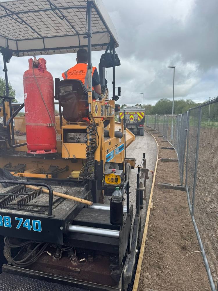 An RM Cleary operator driving the paver, with a delivery lorry waiting ahead.