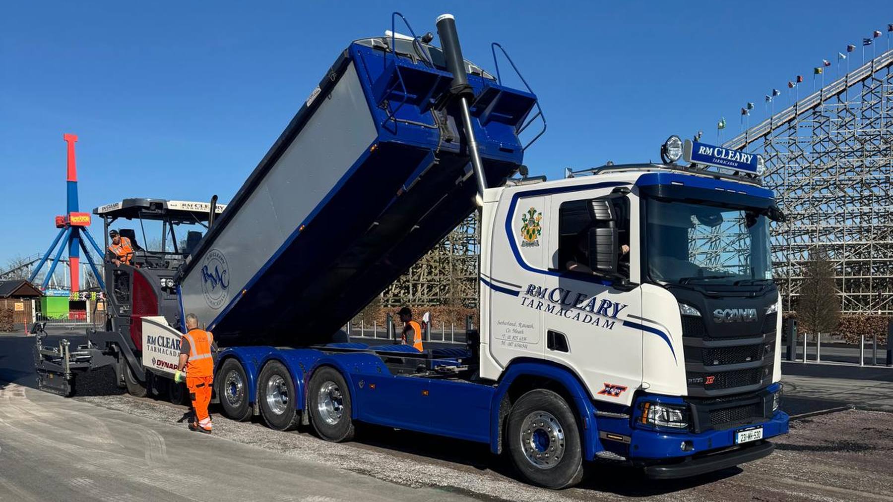 A Scania tipper feeding the paver on a car park resurfacing job under clear blue sky.