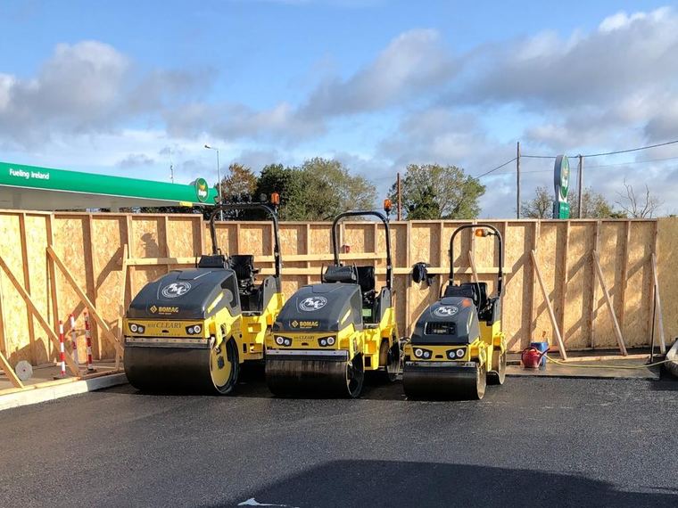 Three yellow RM Cleary BOMAG tandem rollers parked in a row on freshly laid asphalt at a filling station forecourt.