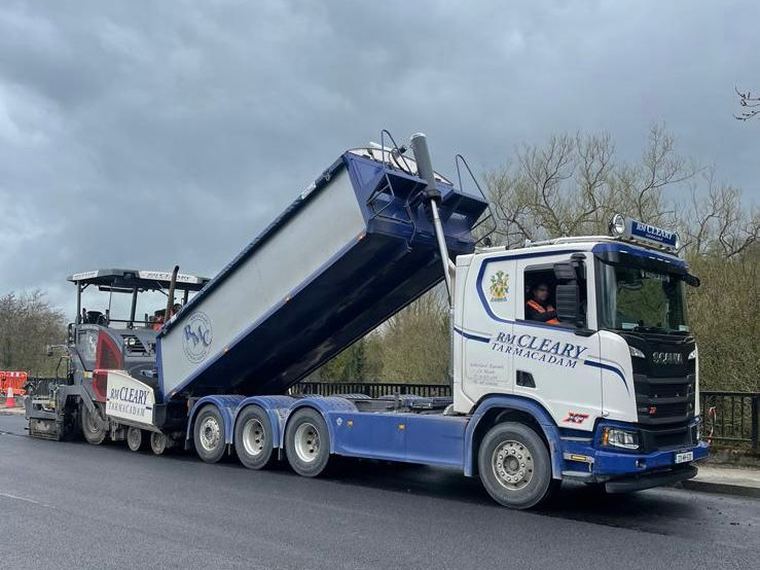 An RM Cleary Scania R530 V8 tipper with the body raised, discharging asphalt straight into the paver on a road job.