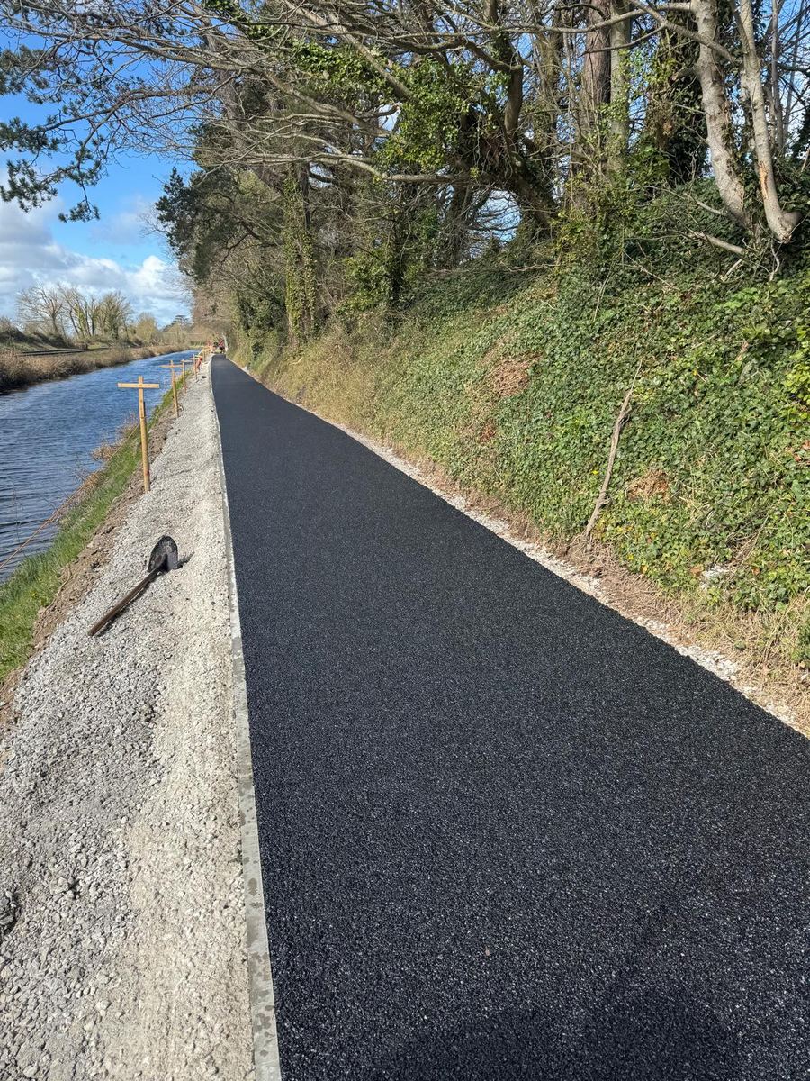 A finished asphalt greenway path running alongside the Royal Canal under a blue sky, kerbed on the canal side with timber marker stakes along the bank.