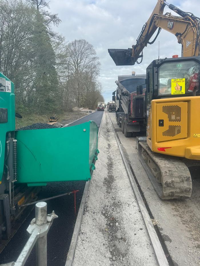 A tipper lorry feeding the paver on the canal bank, with an excavator alongside and further lorries waiting behind.
