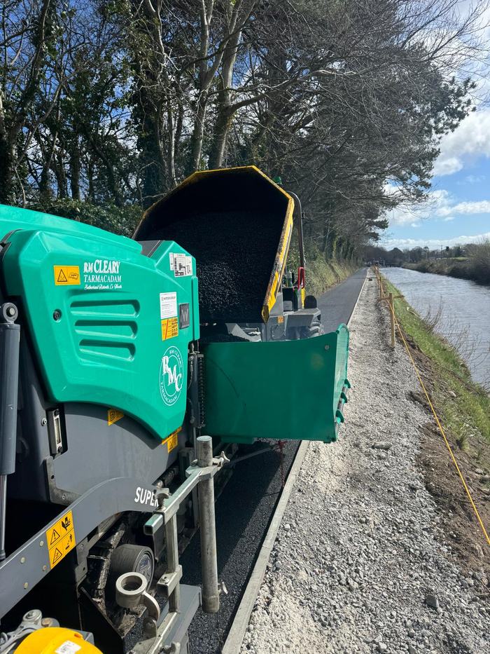 The paver hopper full of hot asphalt beside the canal, with the laid surface running ahead of the machine.