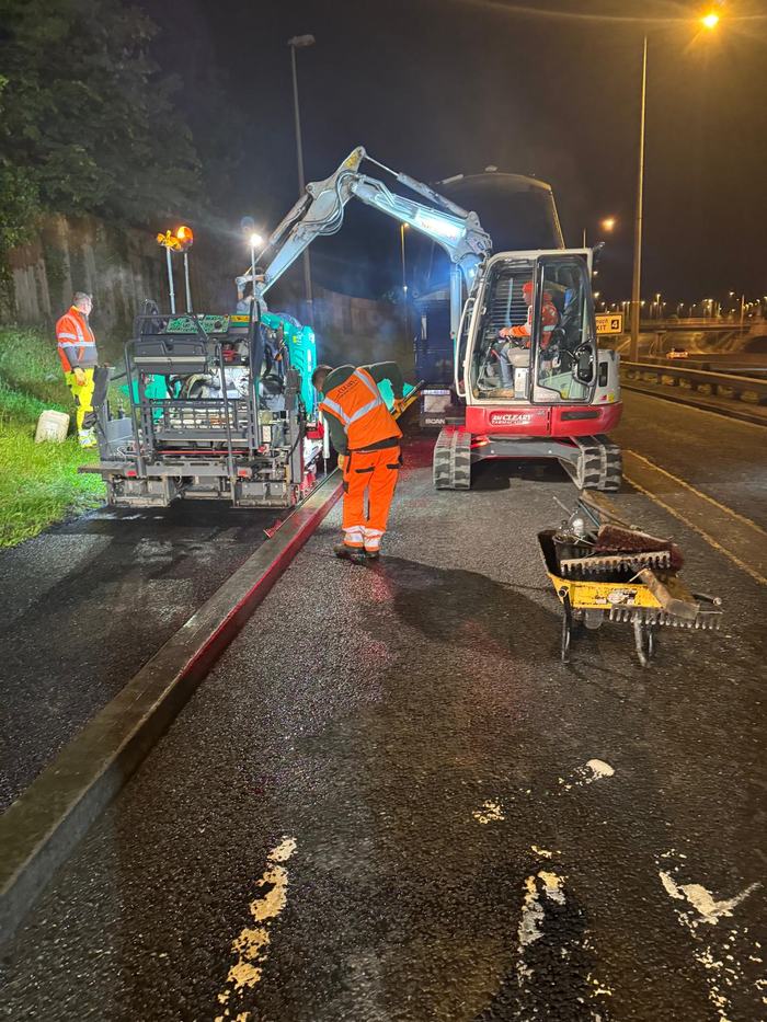 The crew setting the paver up under the site lights beside a motorway slip road, with the excavator waiting behind.