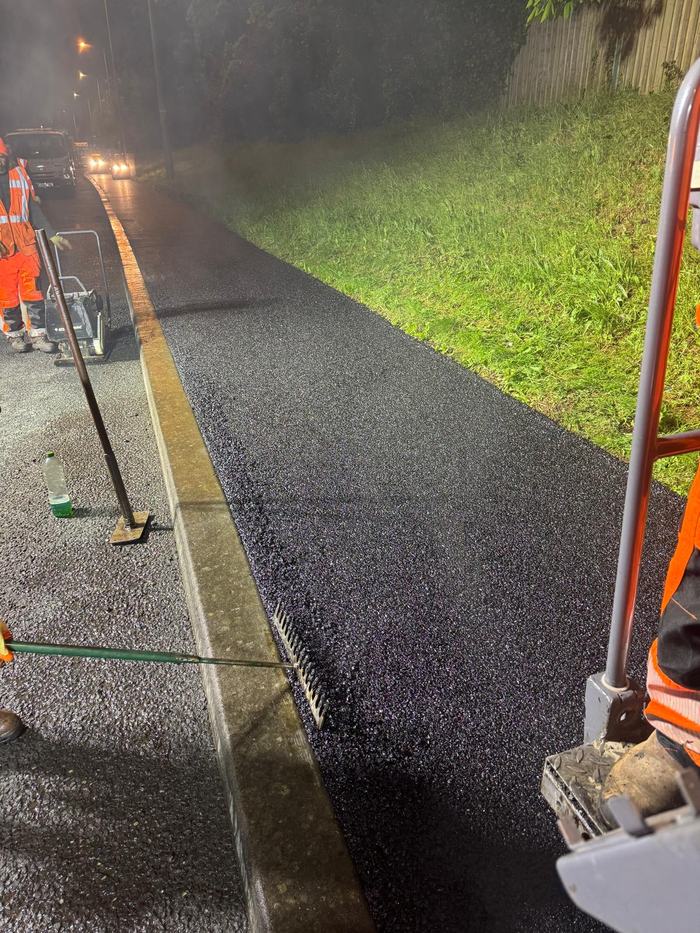 The freshly laid edge running away along the kerb line, still steaming, with the crew and traffic management behind.