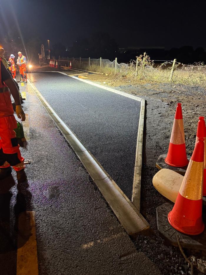 A finished kerbed lane at night, coned off, with the crew clearing up at the far end.