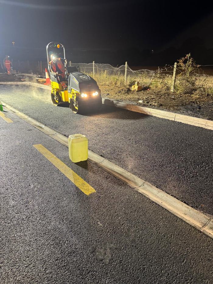 The roller compacting the fresh mat under its own lights, steam lifting off the hot material.