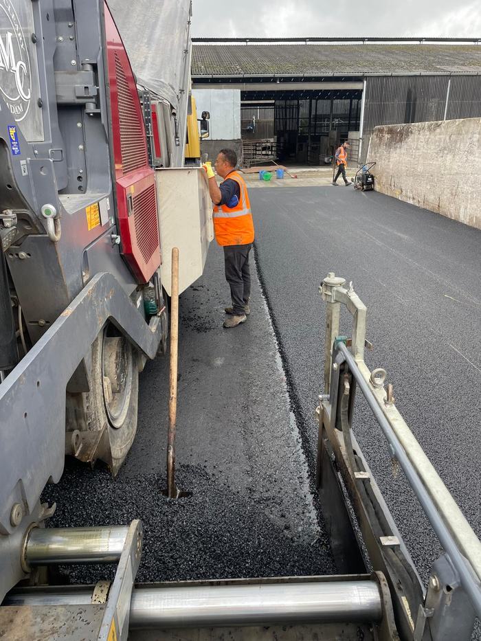 A crew member working the paver hopper with the fresh mat laid out towards the sheds.
